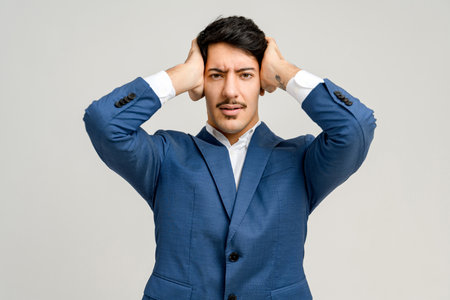 Businessman in a blue suit shows a moment of stress or confusion, gripping his head with both hands, against a plain background, illustrating the challenges and pressures faced in the corporate worldの写真素材