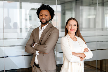 A man and a woman, both dressed in business casual attire, confidently stand with crossed arms in a modern office environment, exuding professionalism and partnershipの写真素材