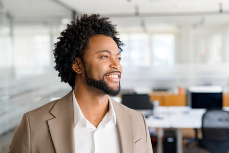 Handsome cheerful businessman in brown suit looking away with a genuine smile, standing in modern office, representing job satisfaction and positive workplace environmentの写真素材