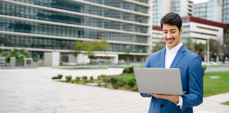 A young Hispanic professional, elegantly dressed in a blue suit, works on his laptop against a backdrop of urban architecture, epitomizing mobile business operations. Business and technology conceptの写真素材