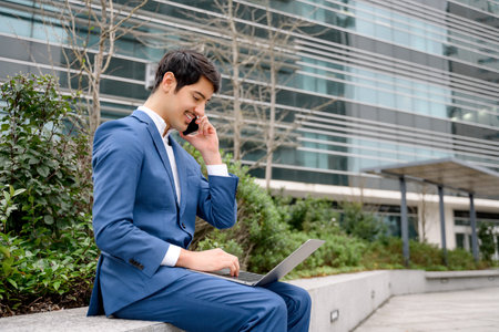 A Hispanic businessman in a stylish blue suit sits outdoors, engrossed in his laptop work while taking a call, blending urban professionalism with the versatility of remote work.の写真素材