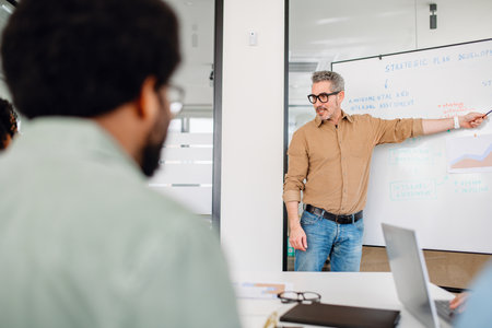 Mature leader mentor enthusiastically points to a strategic plan on a whiteboard, engaging his team...