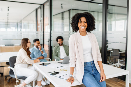 Charming smart Brazilian woman, a female office employee, stands with easy confidence, her team is in the background, embodying a relaxed yet productive meeting atmosphereの写真素材