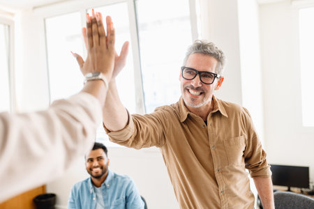 A high-five between colleagues, mature man in focus, emphasizes the exuberant success and positive...