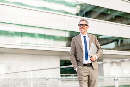 A content senior businessman holding a laptop walks outdoors, his relaxed demeanor and the modern office backdrop illustrating the flexibility of modern executives to work from anywhereの写真素材