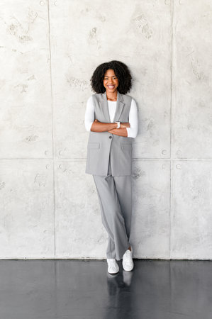 A professional African-American businesswoman stands confidently, arms crossed, with a grey wall on the background, exuding leadership and poiseの写真素材