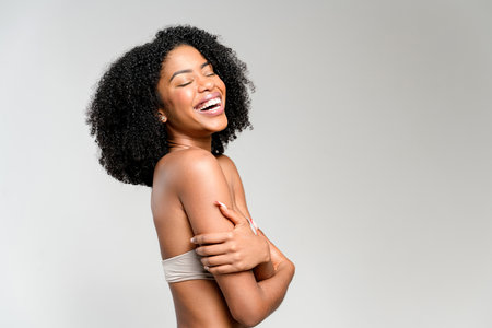 African-American woman laughs heartily, holding her arm across her body, her buoyant curls framing a face filled with mirth, set against a soft grey background that underscores the lighthearted mood.の写真素材