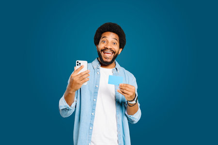 Full of excitement, young Brazilian man looks at the camera while holding a credit card and smartphone after a successful online transaction or receiving good financial news, isolated on blueの写真素材