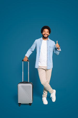 A cheerful young Brazilian man with a captivating smile holds a silver suitcase, giving a thumbs-up against a vibrant blue background, exuding a sense of excitement and anticipation for travelの写真素材