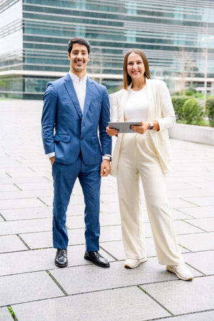A businessman and a businesswoman stand confidently with a tablet, ready to embark on their next venture with the dynamic city backdrop enhancing their forward-thinking attitude.の写真素材