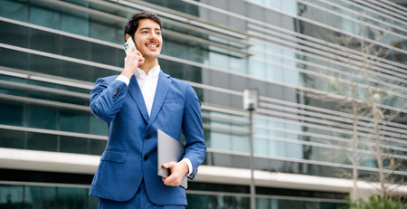 Confident smart Hispanic businessman in a blue suit happily converses on his smartphone while holding a laptop, showcasing flexibility and connectivity in the corporate world.の写真素材