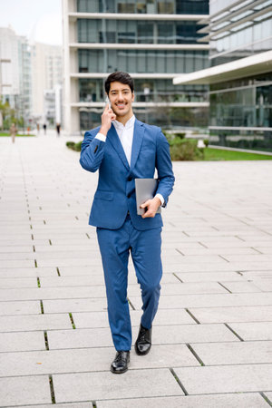 In a bustling cityscape, a young Hispanic businessman smiles as he makes a business call, with a laptop in hand, symbolizing the agility and connectivity of modern professionals.の写真素材