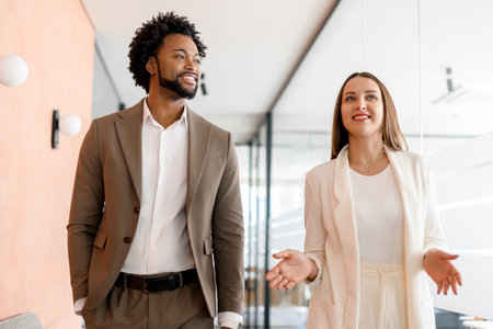 The businessman walks alongside a female colleague in a light and spacious office corridor, suggesting a collaborative work environment. Their interaction implies teamwork and a shared visionの写真素材