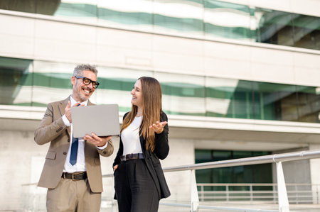 Grey-haired businessman and his female associate engage in a dynamic conversation over a laptop standing outdoors, their expressions reflecting a successful and positive exchange of ideasの写真素材
