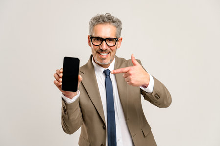 With a charismatic smile, a grey-haired businessman points to a smartphone with an empty screen, suggesting a place for advertising, copy space. Man in suit paying attention on pone display, mock upの写真素材