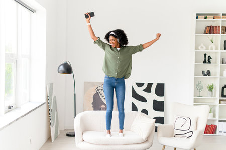 African-American woman celebrates with arms wide open, standing on a sofa in a bright, modern room, using her smartphone to capture the momentの写真素材