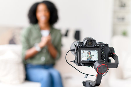 An African-American woman with a radiant smile is seated on a couch, interacting with a camera on a gimbal, creating content in a modern living room with soft natural light, focus on the cameraの写真素材