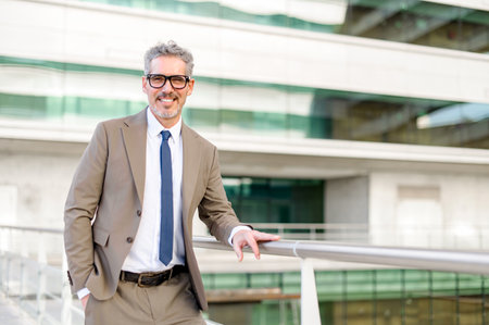 A mature grey-haired handsome businessman is standing at the office balcony and looking at the camera. His engaging smile and direct gaze suggest accessibility and openness.の写真素材