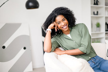 With a hand resting on her cheek, an African-American woman showcases a relaxed and thoughtful expression while sitting in a modern living room, reflecting a serene moment of domestic bliss.の写真素材