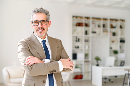 A charismatic senior entrepreneur stands in a modern office space, arms crossed, smiling confidently at the camera, embodying a blend of experience and approachability, focus on leadership and successの写真素材