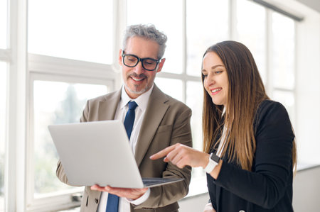 Young female professional points to screen of a laptop while a senior businessman with a jovial expression looks on, indicative of a collaborative and educational moment in a light-filled office spaceの写真素材