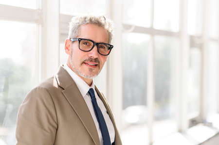 A poised, mature businessman with grey hair and a warm smile stands confidently in a well-lit office, his sharp suit and glasses speaking of wisdom and modern professionalism.の写真素材