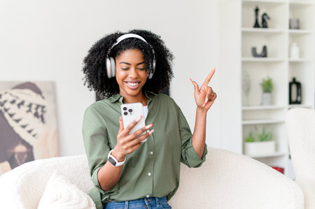 A radiant African-American woman enjoys her music, wearing white headphones and holding a smartphone, her joy palpable in her comfortable home setting. She embodies leisure and connectivityの写真素材