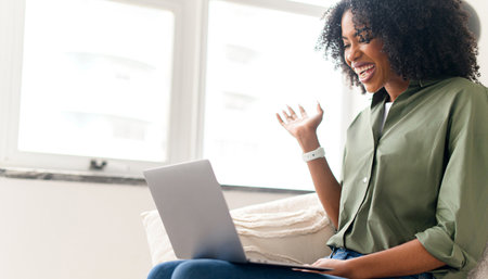 A cheerful African-American woman engages in a video call from her cozy living room, her animated expression suggesting an upbeat conversation. Work-from-home concept.の写真素材