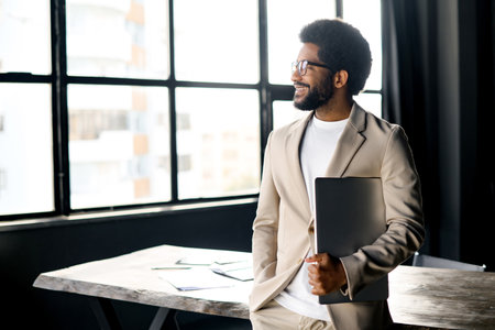Brazilian businessman looks confidently while holding a laptop, standing beside office desk, signifying versatility and mobility in workplace. His engaging smile casts a relaxed yet professional auraの写真素材