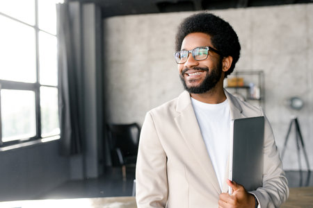 Brazilian businessman holds a laptop, his content smile and casual stance in office reflecting a comfortable command of technology and business. Inspired male entrepreneur full of ideas looks asideの写真素材