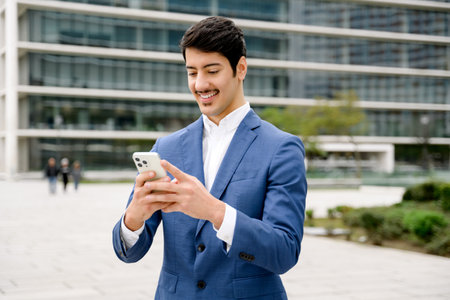 A young Hispanic entrepreneur engages with his smartphone, his expression one of pleasant focus, against a bustling city backdrop that underscores a narrative of connectivity in the digital age.の写真素材