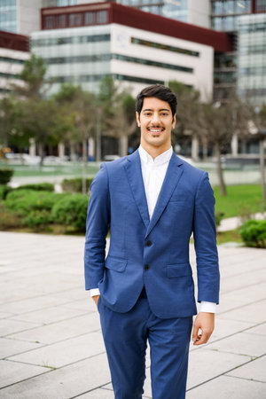 A poised Hispanic businessman presents a friendly demeanor with a gentle smile, standing outdoors in front of a blurred cityscape that hints at a bustling corporate life.の写真素材