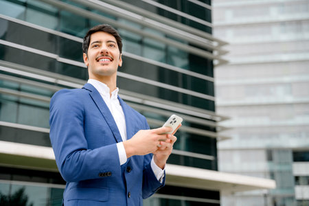 A confident Hispanic businessman holds a smartphone, looking optimistically towards the future against a backdrop of modern architecture. Concept of connectivity and ambition in the corporate world.の写真素材