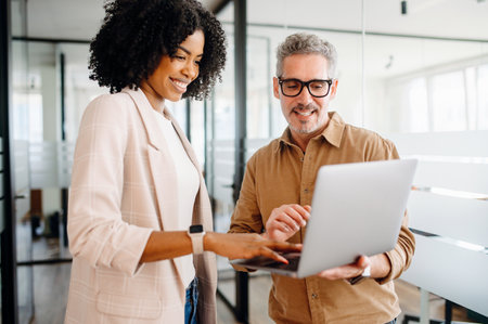 A businesswoman and her mature male colleague explore ideas on a laptop, standing in an office scene that. Image combines expertise and modern tech, blend of wisdom and current technology trendsの写真素材