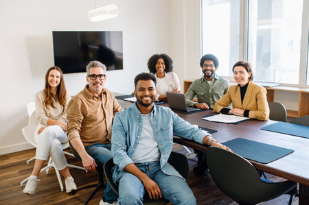 A group of professionals in a modern office setting showcases a cheerful team spirit, with some seated and one standing, all facing the camera with friendly smiles, embodying a productive workplaceの写真素材