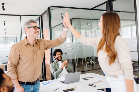 The air is charged with enthusiasm as colleagues share a high-five - one mature experienced mentor man and a young woman, both smiling broadly. The diverse work team applauds, celebrating successの写真素材