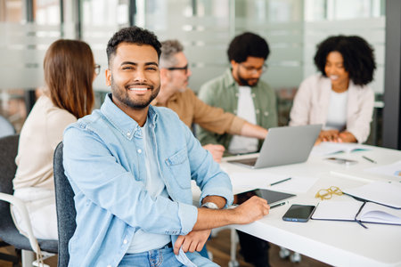 A young man in a blue shirt smiles confidently at the camera, with his colleagues engaged in a discussion in the background, showcasing a harmonious team in a productive settingの写真素材