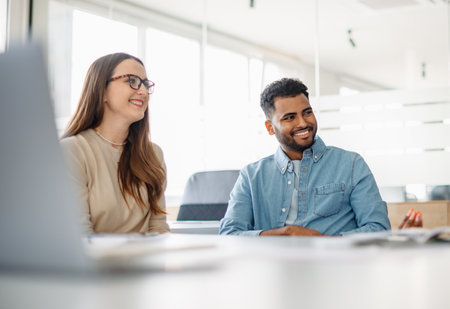 A young woman and a man in a denim shirt are laughing while engaged in a friendly business conversation in a bright, modern office spaceの写真素材