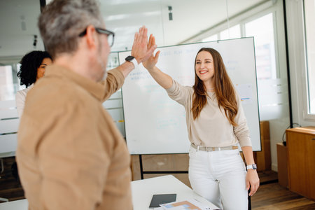 A diverse group of colleagues engages in a moment of team spirit, exchanging high-fives and smiles in a corporate setting. Business success, collaboration, and motivation at their best.の写真素材