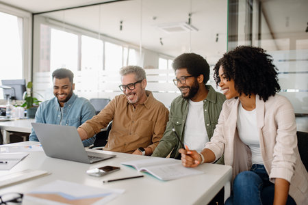 A diverse group of professionals laughs together while working on a laptop in a modern office. The team is engaged in a business discussion, showcasing positive teamwork and collaboration.の写真素材