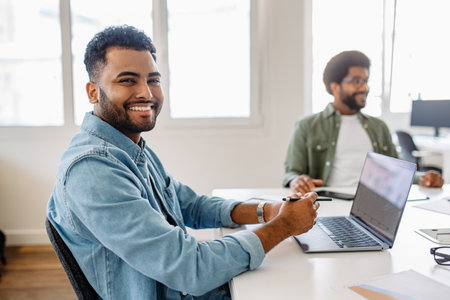 A smiling businessman in a denim shirt working on his laptop in a modern office. His confident and positive demeanor suggests productivity and job satisfaction in a collaborative workspace.の写真素材