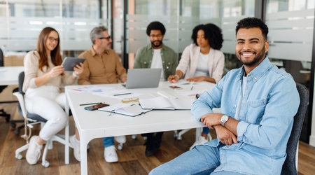 Young professional in denim shirt smiling confidently while sitting at a business meeting with a diverse team in a modern office space.の写真素材