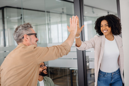 A smiling businesswoman and a senior colleague share a high-five in a collaborative office space. Teamwork, motivation, success, and strong professional relationships in a corporate environment.の写真素材