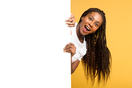 A playful African American woman with curly braids leans behind a blank white panel, laughing joyfully against a bright yellow background. Ideal for modern advertising, branding, and digital marketingの写真素材