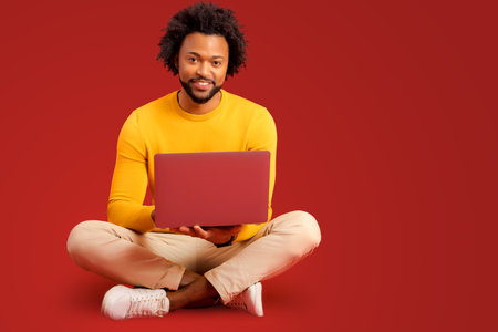 Portrait of happy young bearded man holding and using laptop computer isolated on red background. Male office employee typing, answering email, sittingの写真素材