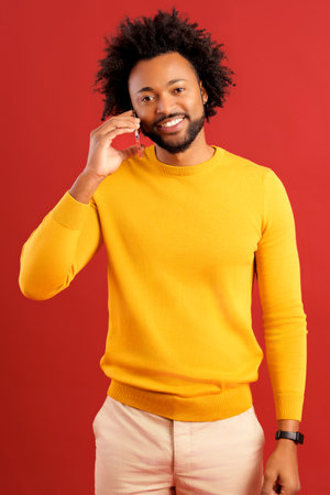 African American curly young man talking by smartphone with empty screen and laughing isolated on red background, overjoyed guy in yellow pullover has pleasant phone call, chattingの写真素材