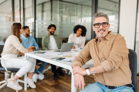 A senior business professional sits at a white office desk, looking directly at the camera with confidence. His diverse team is engaged in a brainstorming session in the backgroundの写真素材