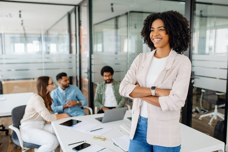 A young female professional stands confidently in a modern office, smiling with her arms crossed. In the background, a diverse team is engaged in a business discussion, collaborating on a project.の写真素材