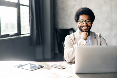 Smiling young office worker at his laptop, displaying an air of casual professionalism in a workspace. His relaxed posture resonate with the flexibility and ease of modern entrepreneurial environmentsの写真素材