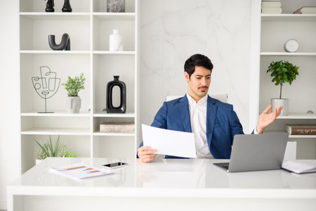 A Hispanic businessman is captured in a moment of frustration or surprise, hand raised in a questioning gesture while reviewing a document at the office desk, his laptop open beside him.の写真素材